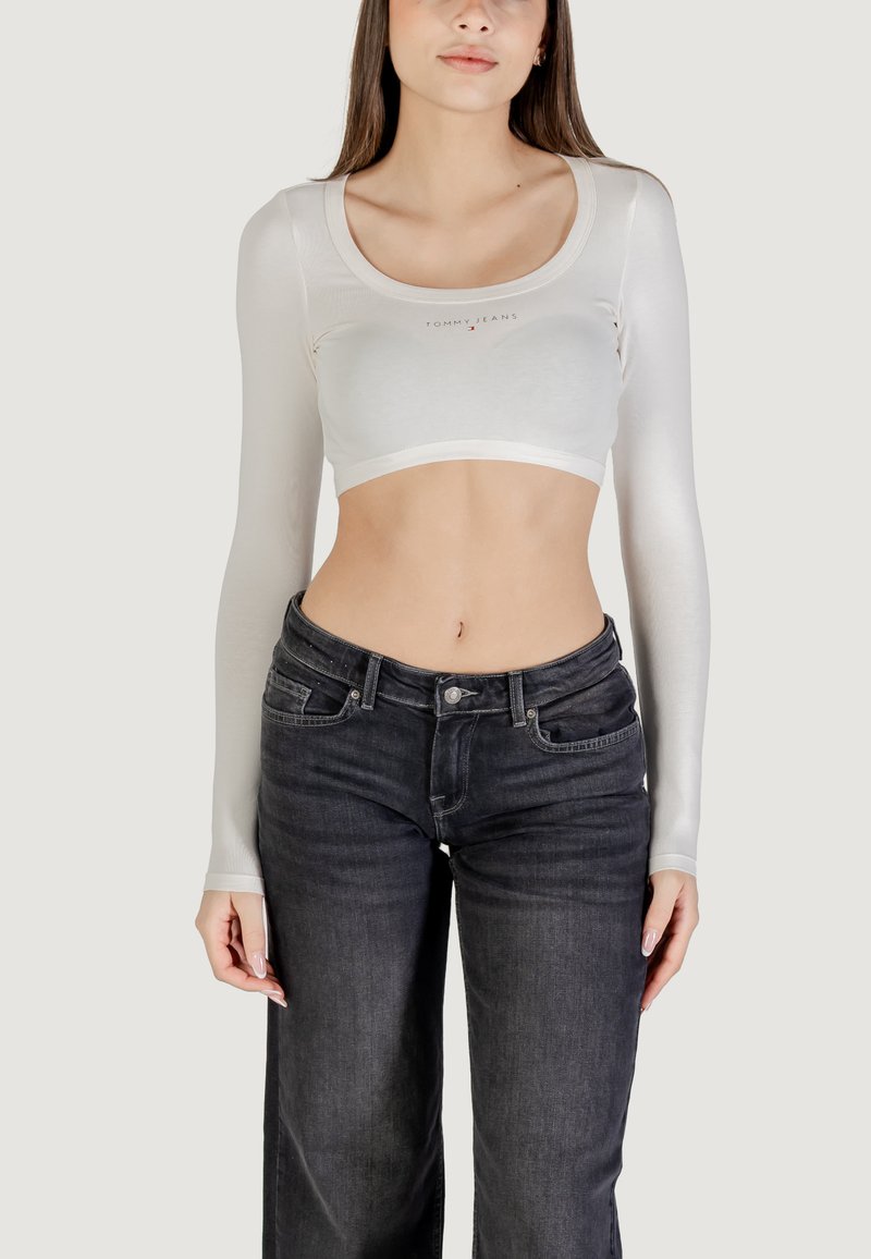 Biały krótki top z długimi rękawami, okrągłym dekoltem i subtelnym detalem z logo na piersi, w połączeniu z ciemnymi jeansami denimowymi. Gładka faktura.