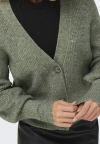 Grön stickad kofta med V-ringning, långa ärmar, en knappknäppning och ribbade muddar, kombinerad med en svart topp under.