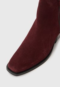 Stivaletto in suede rosso scuro con punta quadrata e suola nera su sfondo bianco.