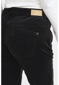 Fekete kordbársony nadrág texturált, bordázott felülettel. Jellemzői között szerepel bőr címke a derékrészen és két hátsó zseb varrási részletekkel.