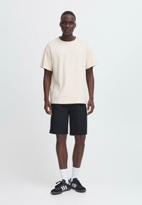 T-shirt en coton beige à manches courtes, associé à un short noir jusqu'aux genoux ; baskets noires avec des rayures blanches, chaussettes blanches.