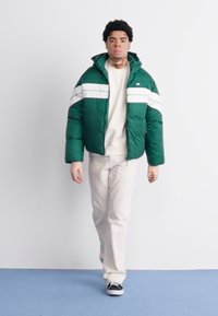 Zelená puferová bunda s bielymi horizontálnymi pruhmi, skombinovaná s béžovým sveterom a pruhovanými nohavicami. Čierne tenisky dotvárajú celý outfit.