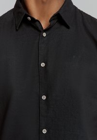 Zwarte button-up shirt van een gestructureerde stof met een klassieke kraag, voorzien van zilverkleurige knopen en een subtiele logo op de borst.