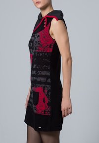 Kvinna i en ärmlös svart klänning med röda och grå abstrakta mönster, framficka och knappad krage mot en enkel grå bakgrund.