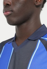 Πόλο μπλούζα με σχέδιο σε ναυτικό μπλε και γαλάζιο, με κάθετες ρίγες, V-λαιμόκοψη και υφασμάτινη υφή για μια δομημένη εμφάνιση.