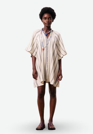 Femme debout portant une robe courte et ample crème avec des rayures noires, des colliers superposés de perles bleues, et des sandales marron à enfiler.