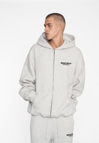 Sudadera con cremallera de color gris claro, hecha de un tejido suave y con un corte holgado. Cuenta con una capucha ajustable y el texto negro "MULTIPLY APPAREL" en el pecho.