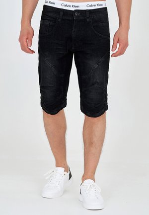 Schwarze Denim-Shorts mit glatter Textur, im enganliegenden Design, knielang geschnitten und mit charakteristischer Taschenverarbeitung. Getragen mit weißen Sneakers.