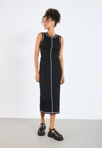 Black ribbed tank dress with a fitted silhouette, featuring white contrast stitching down the front and a knee-length hemline. Paired with black chunky sandals.