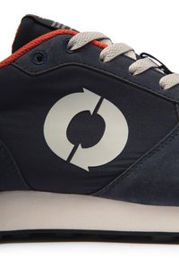 Gros plan d'une basket bleu marine avec un logo flèche circulaire blanc, des lacets blancs, une bordure rouge et une semelle intermédiaire beige.