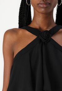 Črni top s halter izrezom, ki se ponaša s prepletenim dizajnom in cvetjem iz blaga na vratu ter mehkim, tekočim materialom.