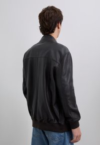 Person, der mit dem Rücken zur Kamera steht, trägt eine schwarze Lederjacke und blaue Jeans vor einem schlichten hellgrauen Hintergrund.