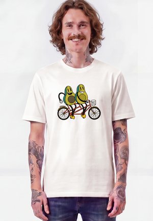 Lächelnder Mann mit schulterlangen Haaren und Tätowierungen, der ein weißes T-Shirt mit dem Aufdruck von zwei Cartoon-Avocados auf einem Tandemfahrrad trägt.