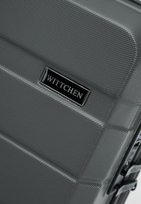 Maleta rígida gris con superficie texturizada, estrías distintivas y una placa de logo negra. Cuenta con cremalleras resistentes y asas laterales.