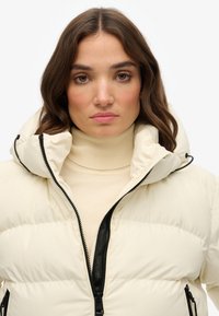 Crème pufferjack met een hoge kraag, zwarte rits en gestructureerde stof. Draagt over een beige coltrui met een zachte, gladde afwerking.
