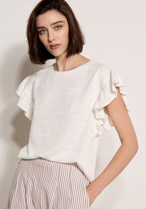 Vrouw met kort bruin haar draagt een wit, gestructureerd blouse met gerimpelde mouwen en beige gestreepte broek met hand in de zak.