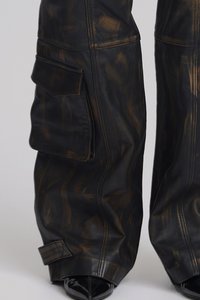 Black and brown stained wide-leg leather pants with large side flap pockets, worn over black pointed-toe shoes on a plain background.