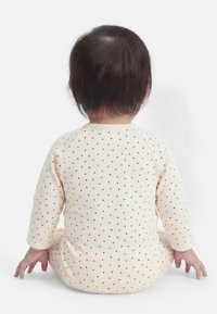 Μωρό με σκούρα μαλλιά που κάθεται αντικρίζοντας προς τα πίσω, φορώντας μπεζ ρούχο με μικρό κόκκινο σχέδιο καρδιάς σε λευκό φόντο.