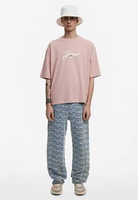 Roza oversized majica s grafičkim dizajnom, uparena s plavim širokim hlačama s uzorkom. Bijela bucket kapa i bež tenisice upotpunjuju izgled.