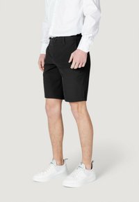 Pantalones cortos negros de tela sintética, con corte recto, presillas para cinturón y bolsillos frontales. Combinados con zapatillas blancas.
