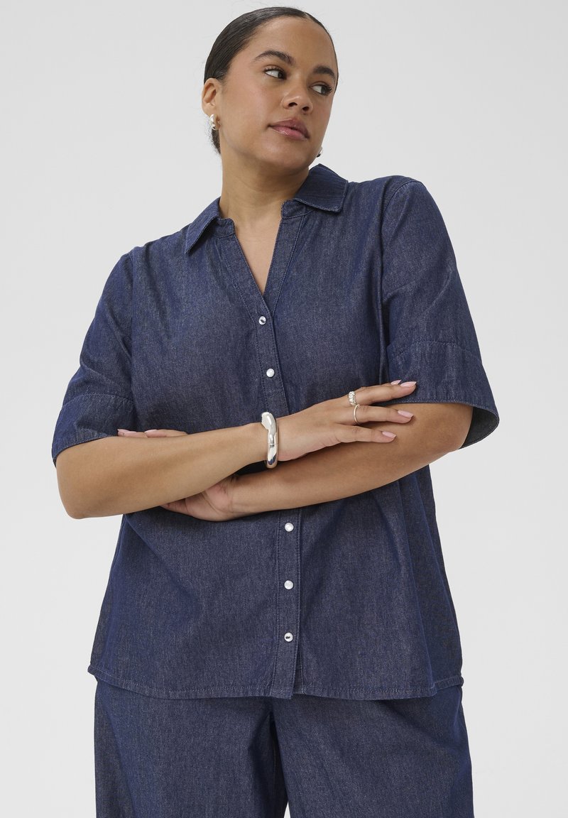 Femme les bras croisés portant une chemise boutonnée à manches courtes bleu foncé et un pantalon assorti, accessoirisée d'un bracelet en argent et de bagues.