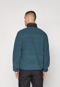 Chaqueta de forro polar en color verde azulado con un cuello alto y acentos en negro, con una superficie texturizada y costuras de contraste en la parte trasera.