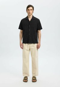 Chemise noire à manches courtes avec un motif en spirale texturé, associée à un pantalon beige à jambes larges et des sandales vertes. Boutons simples sur la chemise.
