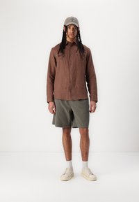 Camisa de lino marrón con botones, pantalones cortos verde oliva, zapatillas beige y una gorra gris claro. Conjunto casual con una textura de tela natural.