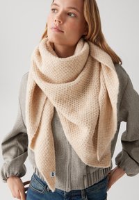 Femme portant une grande écharpe beige texturée enroulée autour de son cou et de son épaule, sur une veste grise zippée et un jean bleu.