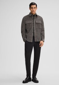 Giacca camicia di corduroy grigia con due tasche frontali, chiusura a bottoni, maniche lunghe e vestibilità rilassata, abbinata a pantaloni neri e scarpe.
