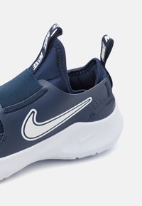 Baskets slip-on bleu marine avec bande élastique, un logo Nike Swoosh blanc contrastant et des accents en tissu texturé, dotées de languettes pour un enfilage facile.