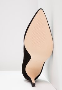 Chaussure à talons hauts en daim noir avec un bout pointu, présentant une semelle beige lisse et une texture antidérapante. Le talon est fin et effilé.