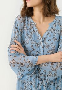 Svetle modré květinové šaty s volnými rukávy, knoflíkovým výstřihem a malými tmavými květinovými vzory po celém povrchu. Měkká, lehká textilie.
