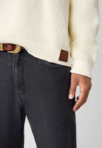 Elfenbeinfarbener Strickpullover mit strukturiertem Muster und gerippten Bündchen, kombiniert mit dunkelblauen Jeans und einem braunen Ledergürtel mit goldener Schnalle.