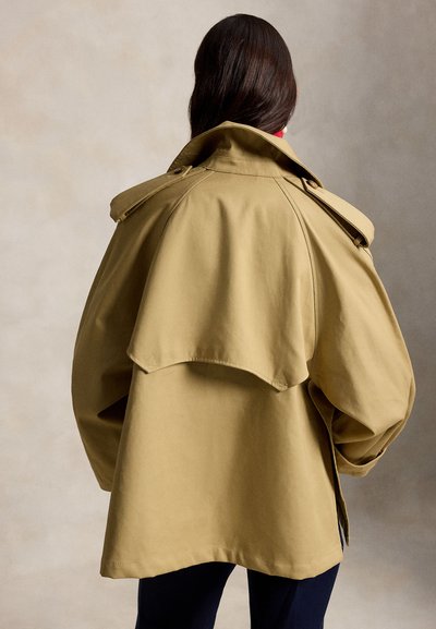 Άτομο με μακριά σκούρα μαλλιά που φοράει φαρδιά καμηλό καμπαρντίνα, εμφανιζόμενο από πίσω με ουδέτερο φόντο.