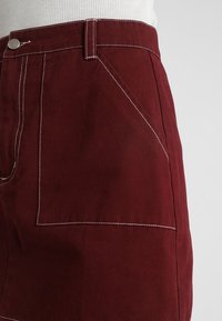 Burgundská sukně z pevných tkanin s bílými švy, s přední kapsou, vysokým pasem a čistým, elegantním střihem.