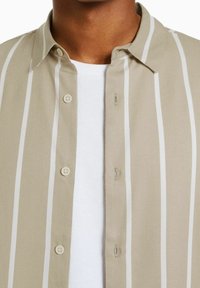 Beige kauluspaita, jossa on valkoisia pystysuoria raitoja, pehmeällä pinnalla, kauluksella ja kahdella nappikiinnityksellä, kerrottu valkoisen t-paidan päälle.