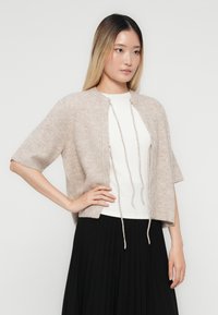 Hellbeige gestrickte Cardigan mit kurzen Ärmeln und rundem Ausschnitt, mit einer Schnürung und einer weichen, strukturierten Oberfläche. Getragen über einem weißen Oberteil.
