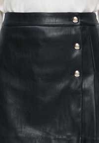 Jupe en cuir noire avec un rabat avant asymétrique fixé par trois boutons décoratifs argentés sur le côté droit.