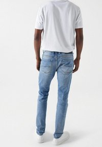 Light blue denim jeans with a slim fit, faded texture, and back pockets featuring subtle stitching. Paired with a plain white t-shirt.