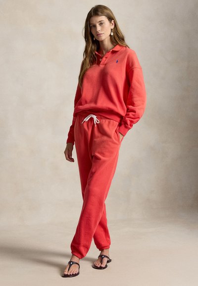 Joven de pie con una camisa polo de manga larga color coral y pantalones deportivos a juego, con chanclas negras y pendientes colgantes.