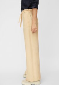 Persona che indossa pantaloni beige a gamba larga con vita elastica e lacci, abbinati a scarpe chunky di un bianco sporco e una maglietta scura con maniche arrotolate.