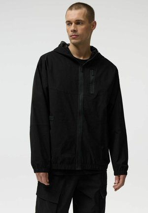 Młody mężczyzna w czarnej, zakładanej na zamek bluzie z kapturem i czarnych spodniach typu cargo, stojący na tle jasnego, jednolitego tła, patrzący w bok.