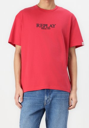 T-shirt en coton rouge à manches courtes, col rond, avec le logo "REPLAY SINCE 1981" en noir. Coupe droite et ajustement décontracté. Porté avec un jean bleu.