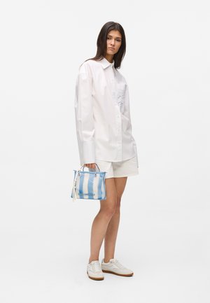 Femme portant une chemise blanche à boutons oversize et un short blanc, tenant un petit sac à main bleu et blanc rayé, debout devant un fond blanc.
