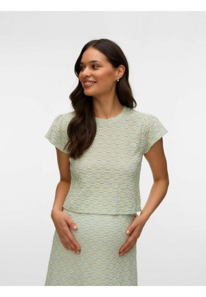 Kvinde med langt mørkt hår iført en lyseblå og gul tekstureret kortærmet top og nederdel, smilende og med hænderne på livet.