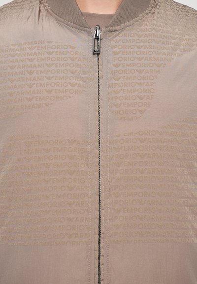 Jachetă bomber de culoare bej deschis, cu un model texturat de logo, guler ribbed și fermoar central. Materialul are un finisaj neted, ușor reflectant.