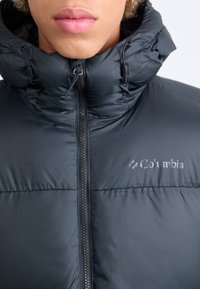 Svart puffjacka med hög krage, med en framdragkedja och justerbara snören. Columbia-logotypen på övre vänstra sidan.