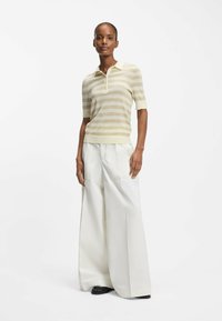 Donna in piedi che indossa una polo a righe beige con maniche corte e pantaloni bianchi a gamba larga, con scarpe nere, su uno sfondo bianco.