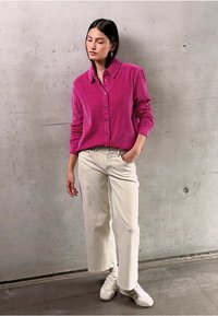 Camicia a bottoni in corda fucsia con colletto, abbinata a pantaloni larghi a vita alta color crema e sneakers bianche, con sfondo un muro di cemento.